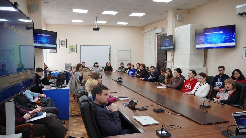 студенти сидять за довгим овальним столом. На стінах висять телевізори