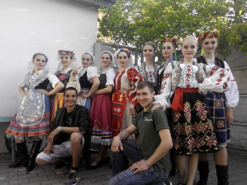 Два хлопці сидять, а за їхніми спинами стоїть дівочий ансамбль у вишиванках