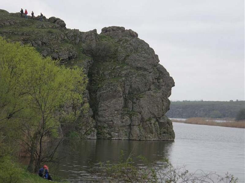 Велика скеля врізалася у воду річки. На вершині скелі люди, у її підніжжя на березі теж сидить людина
