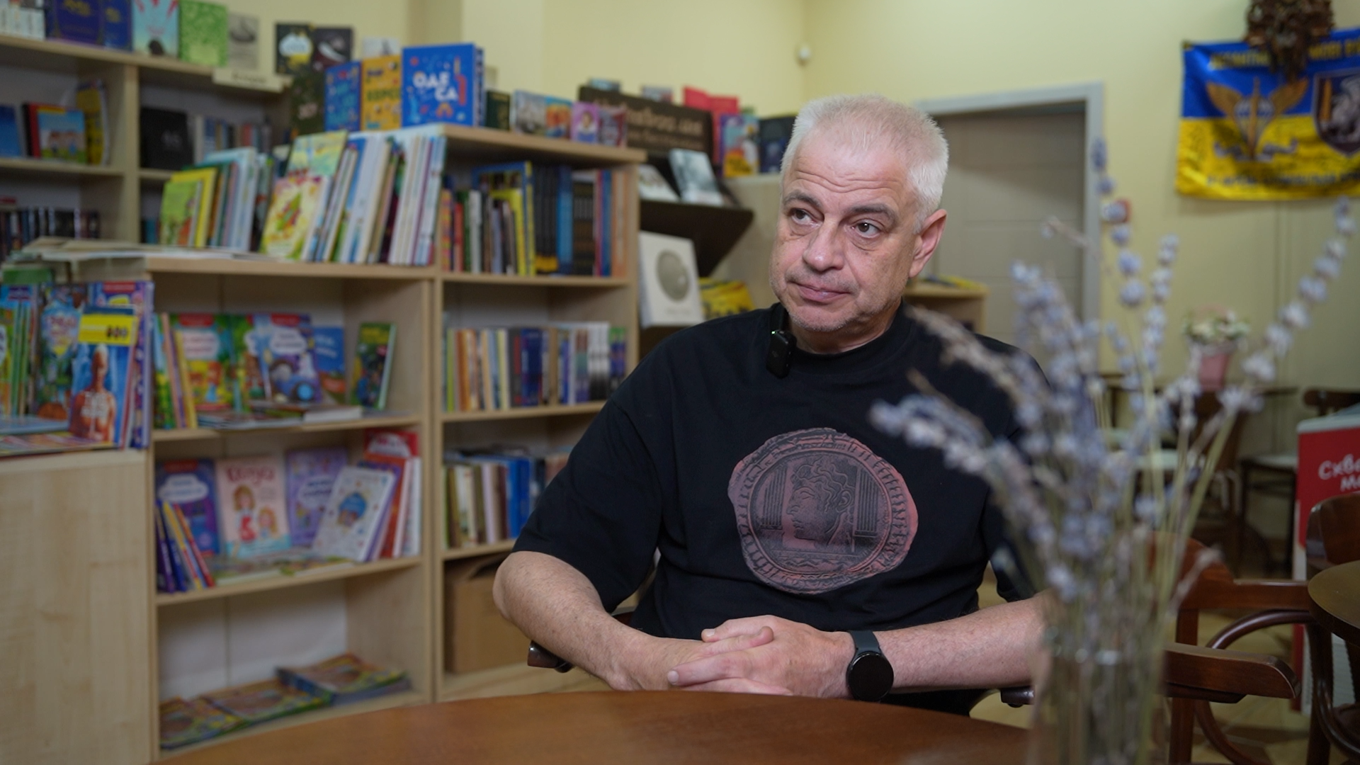 Геннадій Сульдін сидить за столом, за його спиною книжкові полиці
