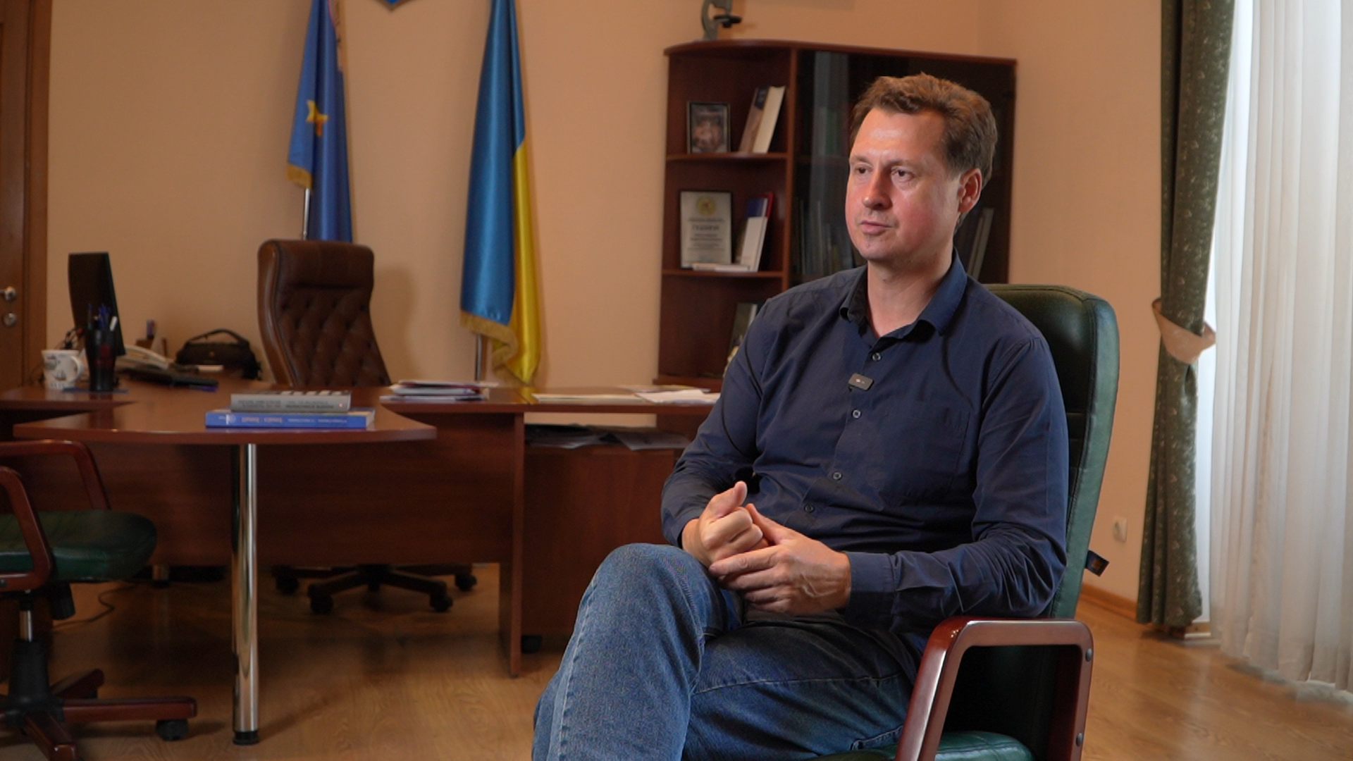 Чоловік сидить у кріслі у центрі красивого кабінету. Біля стіни - прапори України