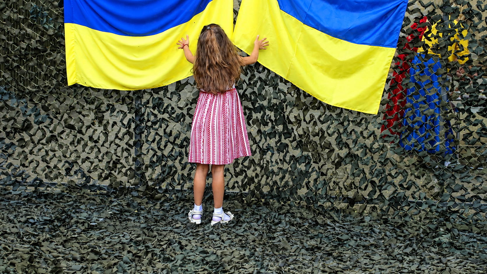 Дівчинка стоїть спиною до камери й торкається двох українських прапорів